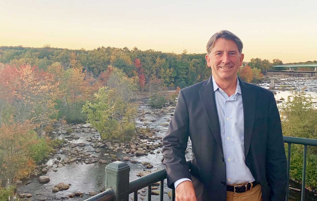 Man in a blazer stands by a railing overlooking a river and autumn trees at sunset, representing ambition and leadership, much like participants of the Goldman Sachs 10,000 Small Businesses program.