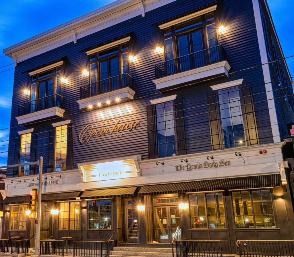 A three-story restaurant with warm lights, dark blue siding, and large windows at dusk&mdash;an inviting spot often featured in local media relations stories.