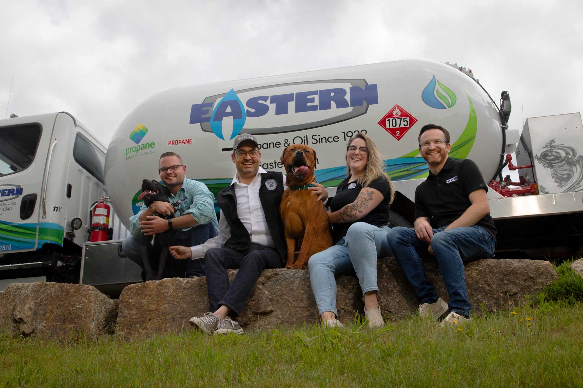 Four people and two dogs sit on rocks in front of an Eastern propane truck, smiling at the camera.