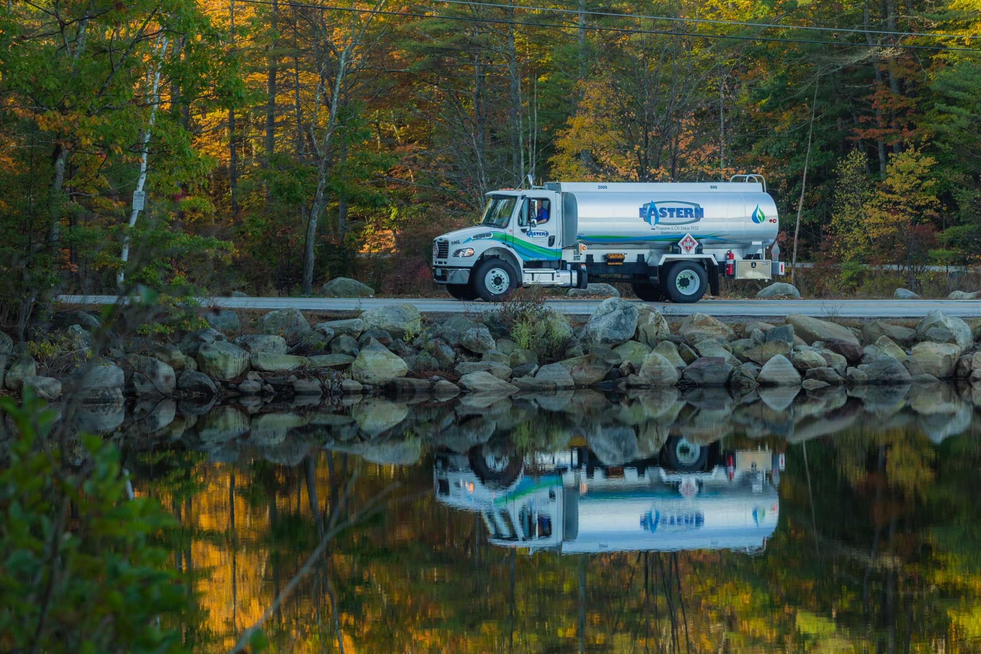 A fuel truck drives past a lake with autumn trees reflected in the calm water.