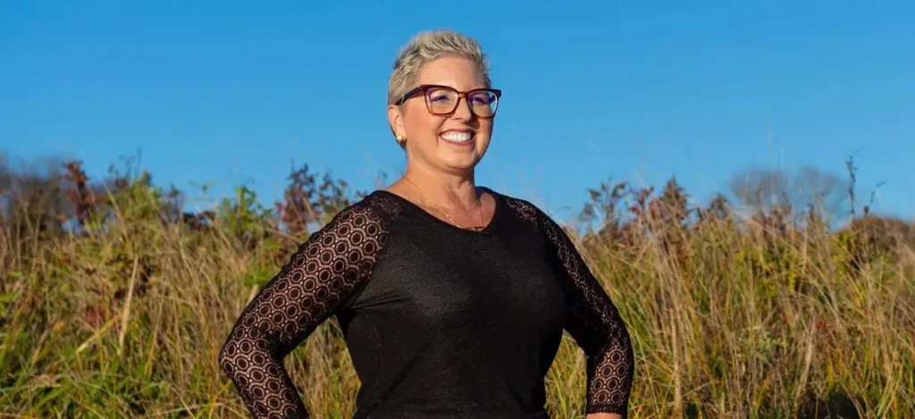 Smiling woman with short gray hair and glasses stands in front of tall grass under a clear blue sky.