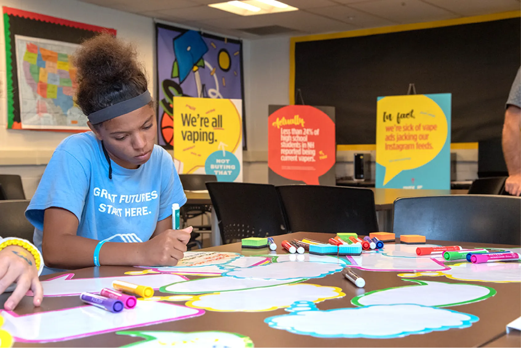 A girl writes on paper clouds at a table covered with markers in a classroom with colorful posters behind her.