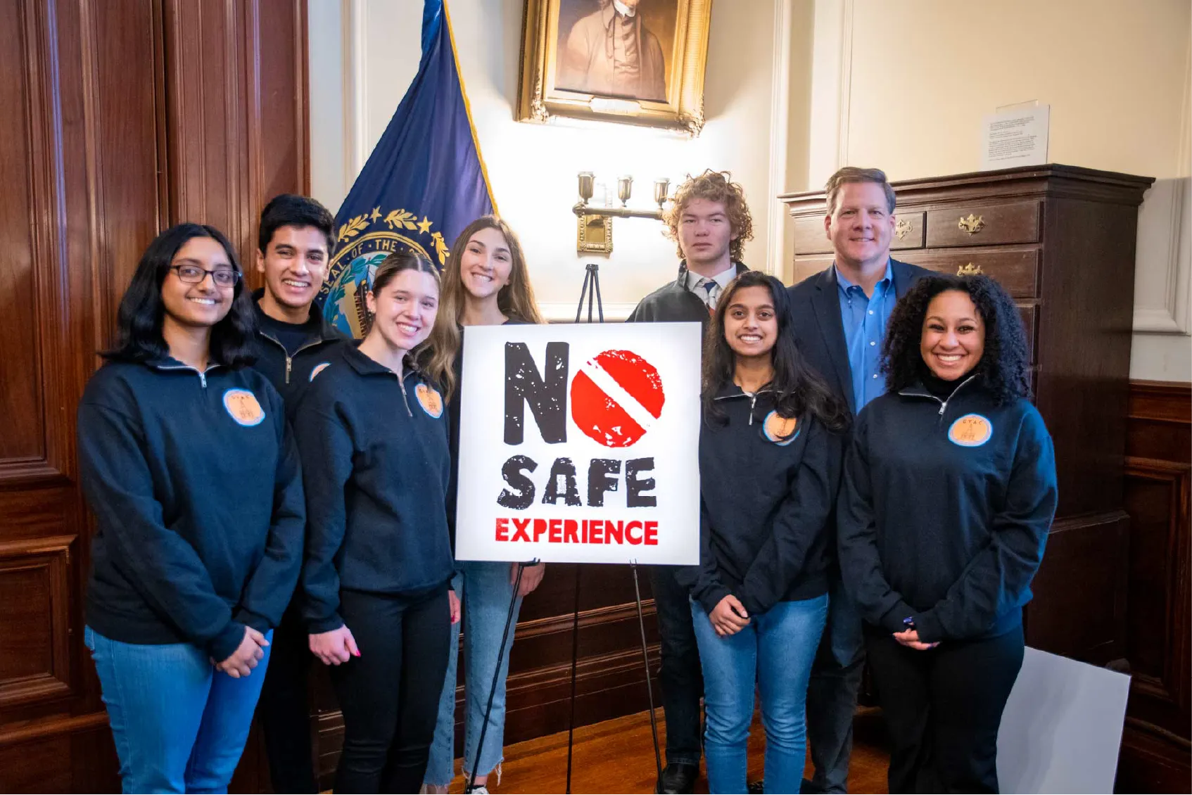 Seven people stand indoors around a sign that reads NO SAFE EXPERIENCE in bold letters.