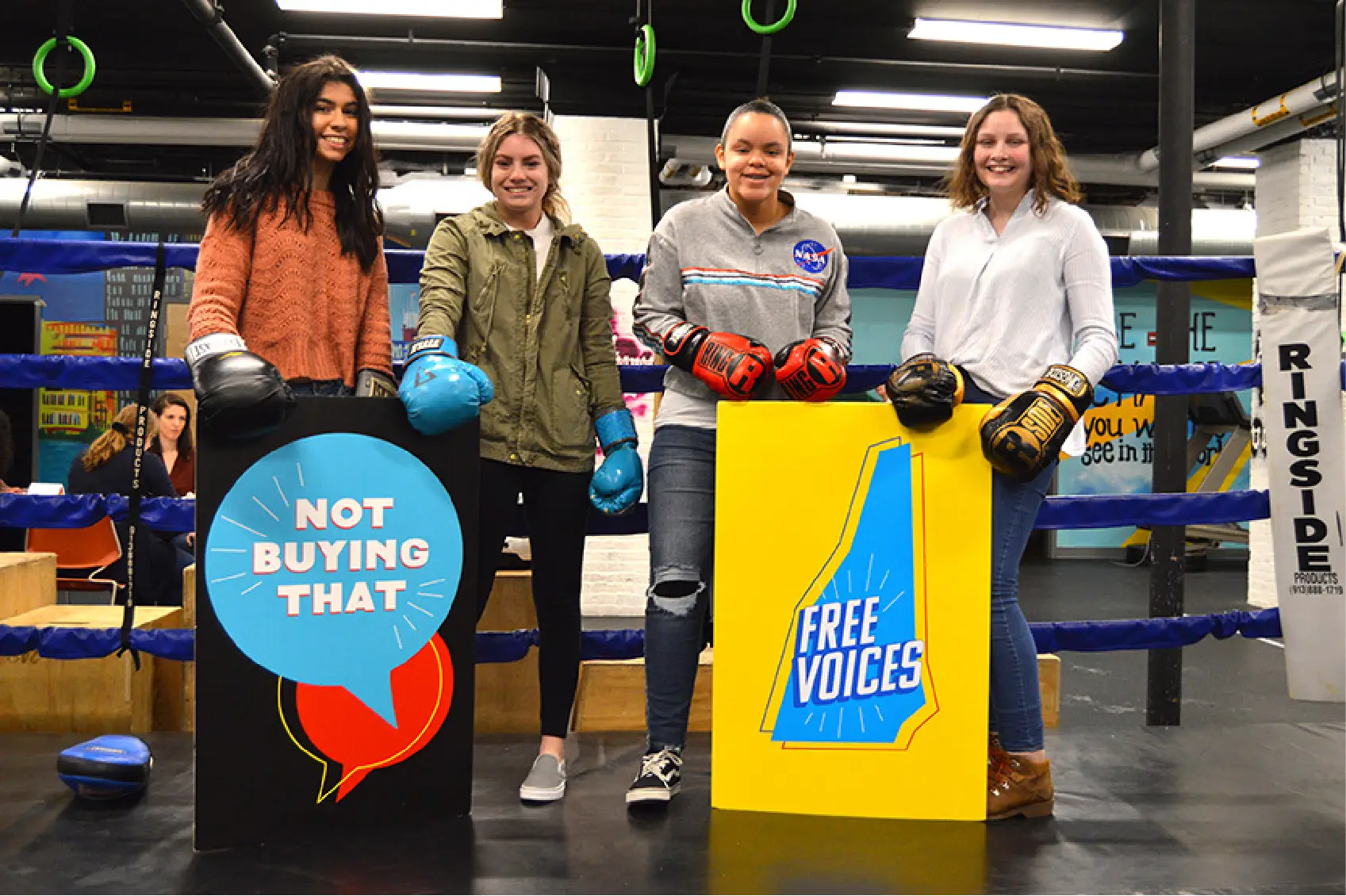 Four young women in a boxing gym, wearing boxing gloves, stand united for Community empowerment as they hold signs reading &ldquo;Not Buying That&rdquo; and &ldquo;Free Voices&rdquo;&mdash;a bold statement against manipulative marketing.