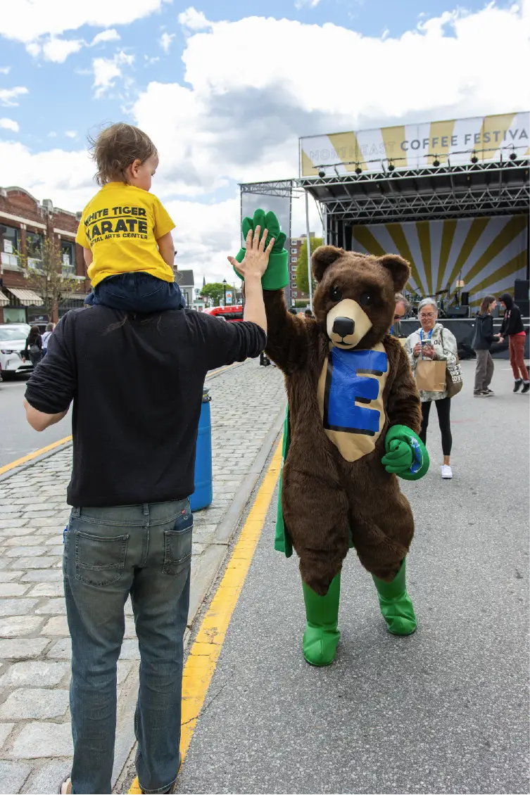 A child on an adult&rsquo;s shoulders high-fives a bear mascot at an outdoor street festival, capturing the spirit of community.