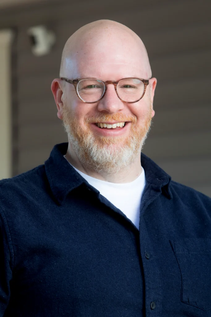 A smiling bald man with glasses and a beard, wearing a navy shirt and white t-shirt, stands outside&mdash;this is Steve Morin.