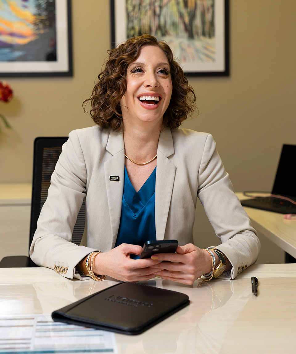 Woman with curly hair smiling, seated at a desk, holding a smartphone, with a folder and pen in front of her&mdash;ready to tackle her next finance project.