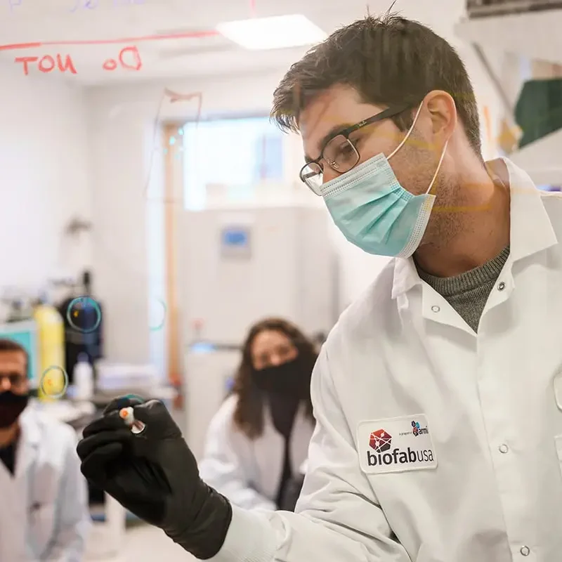 Scientist in a lab coat and mask writes on a transparent board, brainstorming marketing strategies with two colleagues in the background.