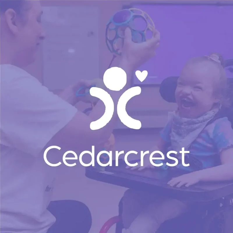 A smiling child in a wheelchair plays with a caregiver under the Cedarcrest logo and name, capturing a heartwarming moment ideal for marketing.