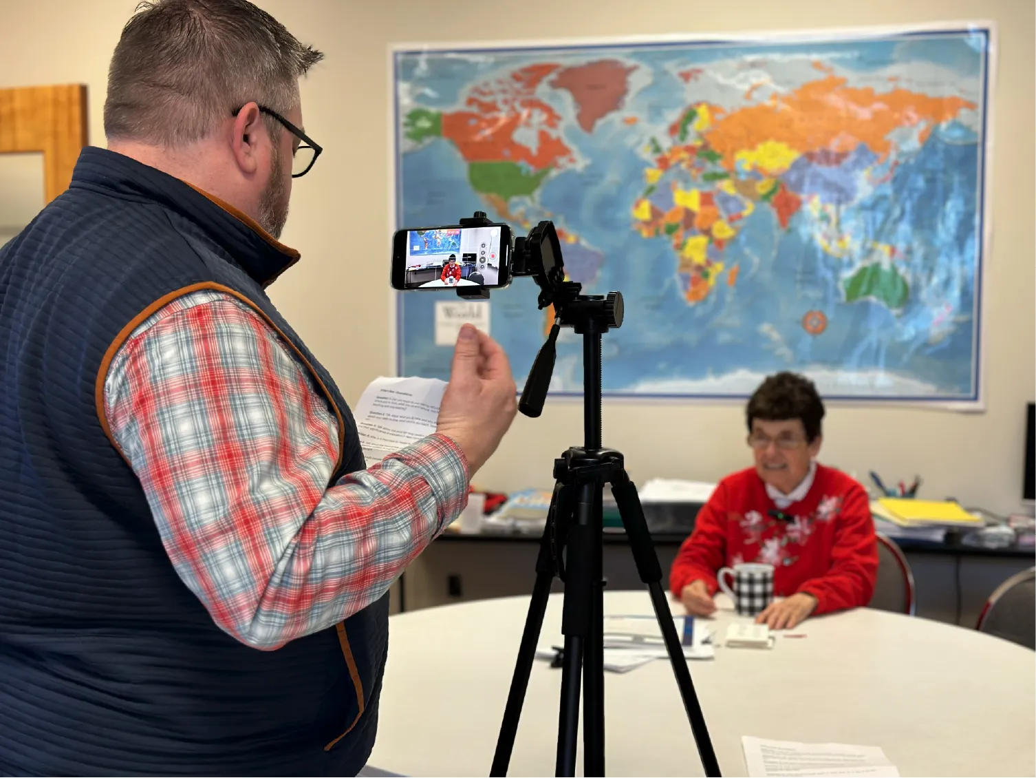 A man records a woman at a table with papers in front of a world map using a smartphone on a tripod.