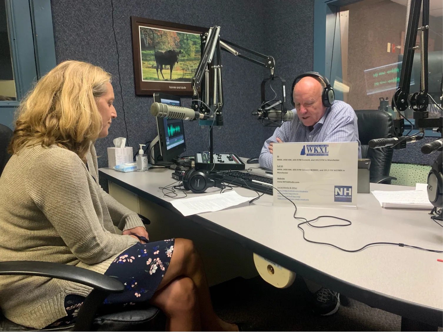 Two people sit in a radio studio at microphones, having a conversation across a table.
