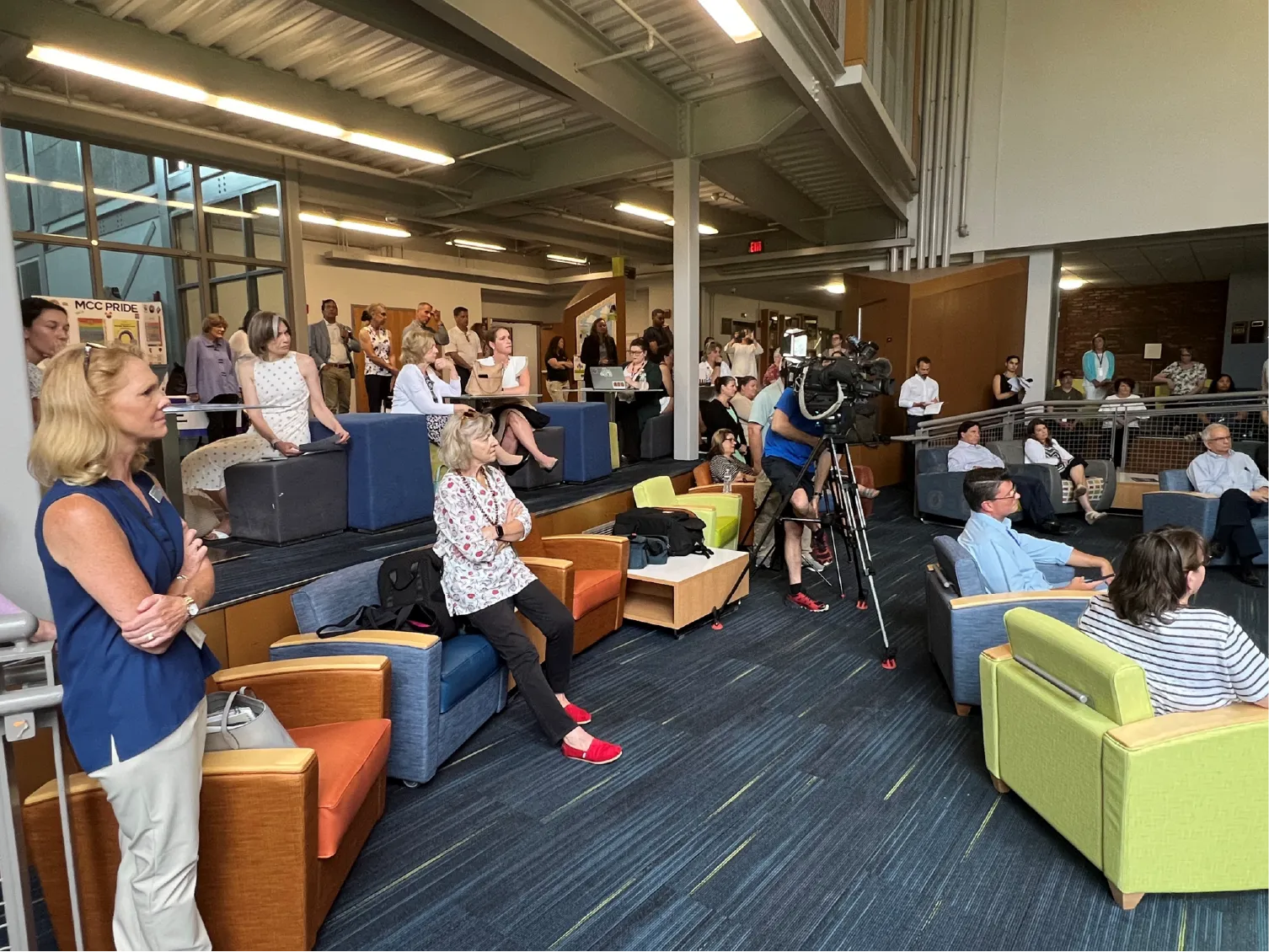 People sitting and standing in a modern lounge area, some watching a speaker, with a camera crew filming.