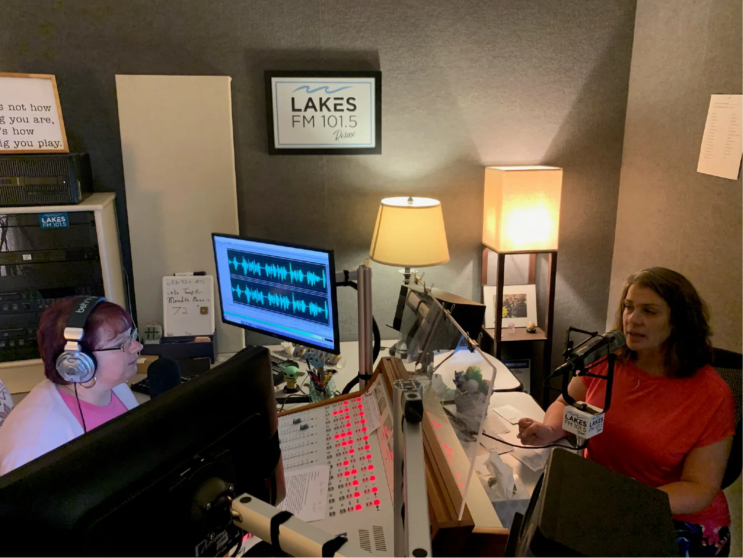 Two women host a radio show in a studio with microphones, computers, and a LAKES FM 101.5 sign.