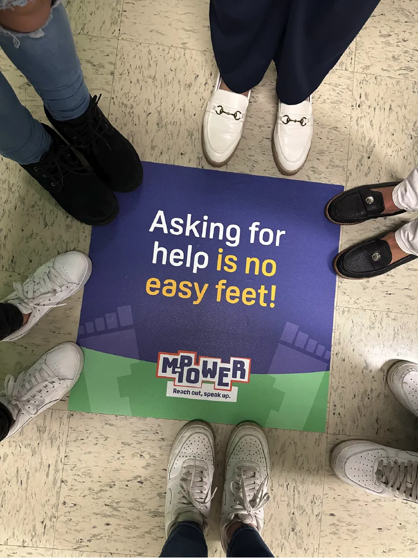 People&rsquo;s feet in a circle around a sign that reads, &ldquo;Asking for help is no easy feet! MPOWER.&rdquo;.