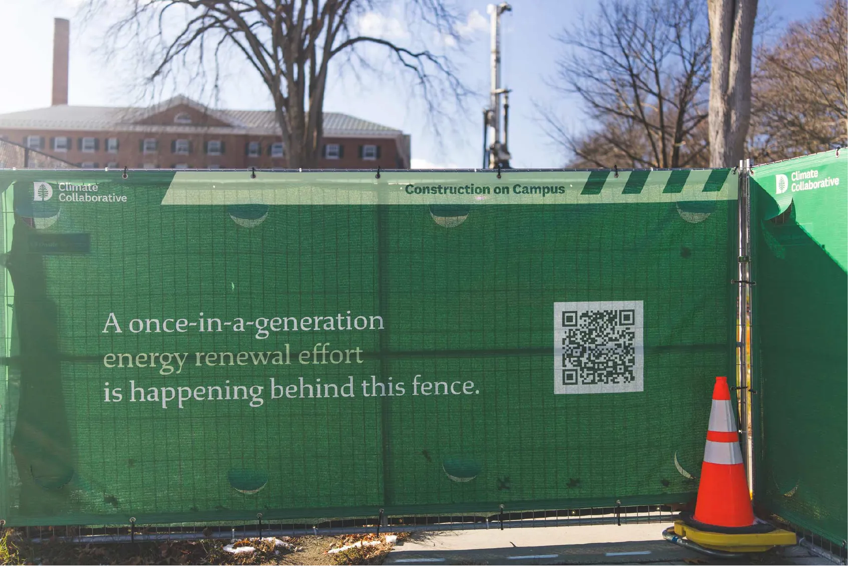 Green fence with sign about an energy renewal effort; QR code and two orange traffic cones in front.