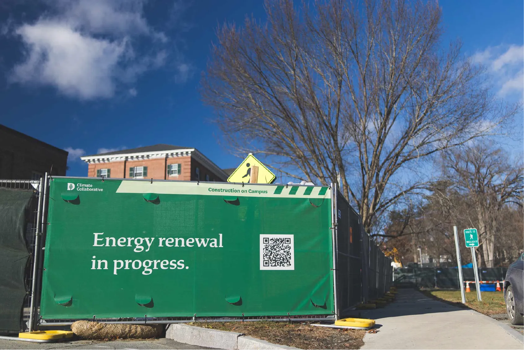 A green construction fence reads, Energy renewal in progress, with a QR code and campus buildings in the background.