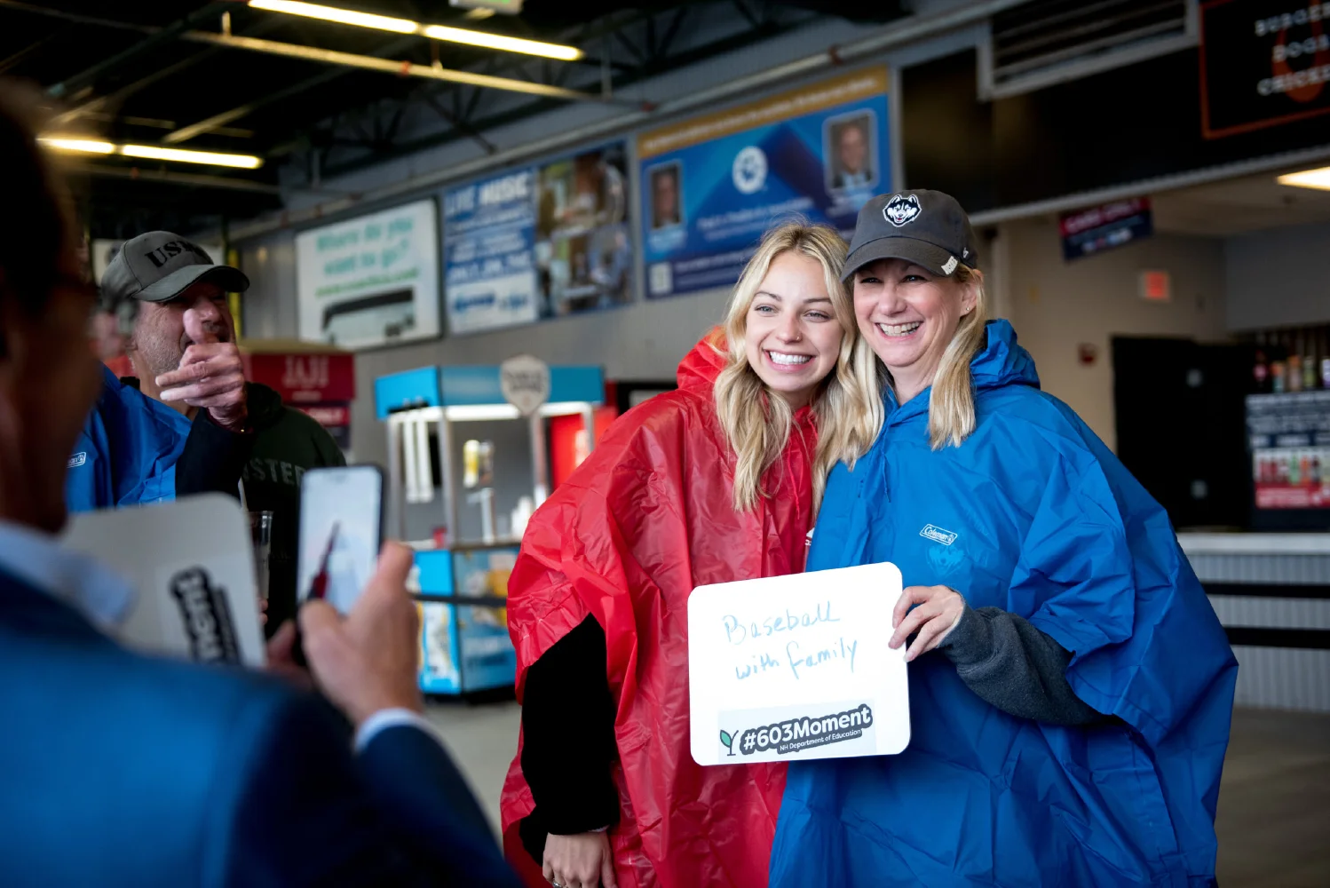 Two women in rain ponchos smile for a photo, holding a "Baseball with family" sign&mdash;a picture-perfect moment that captures memories and the spirit of growth marketing by bringing communities together.
