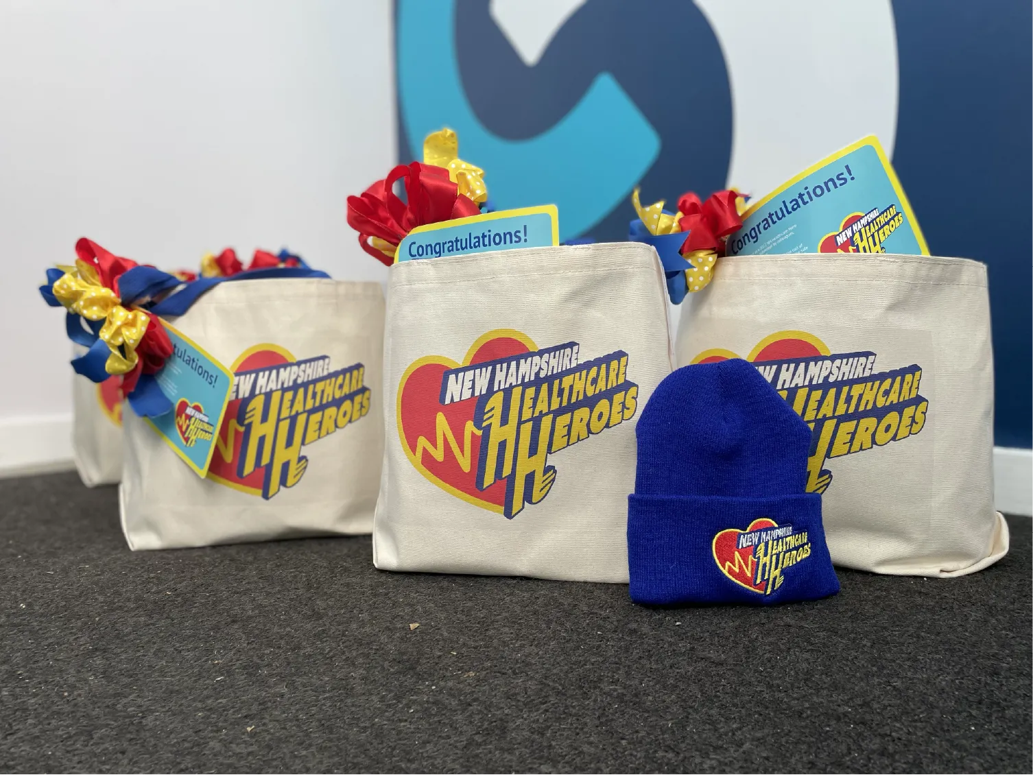 Canvas bags labeled &ldquo;New Hampshire Healthcare Heroes&rdquo; with gifts and a blue knit hat on a carpeted floor.