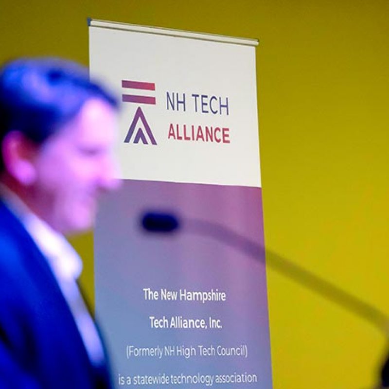 A blurred man speaks near a NH Tech Alliance sign with a yellow background, highlighting the importance of marketing in today&rsquo;s tech-driven world.