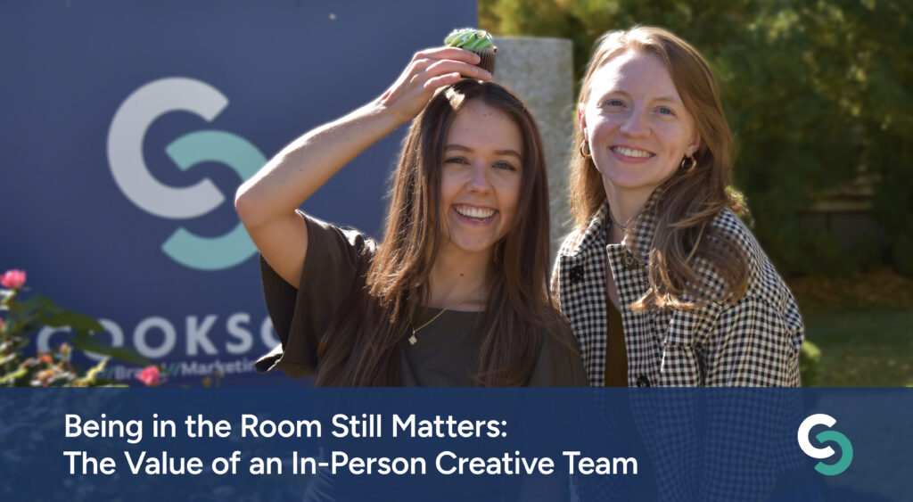 Two women smiling, one balancing a cupcake on her head, in front of a company sign.